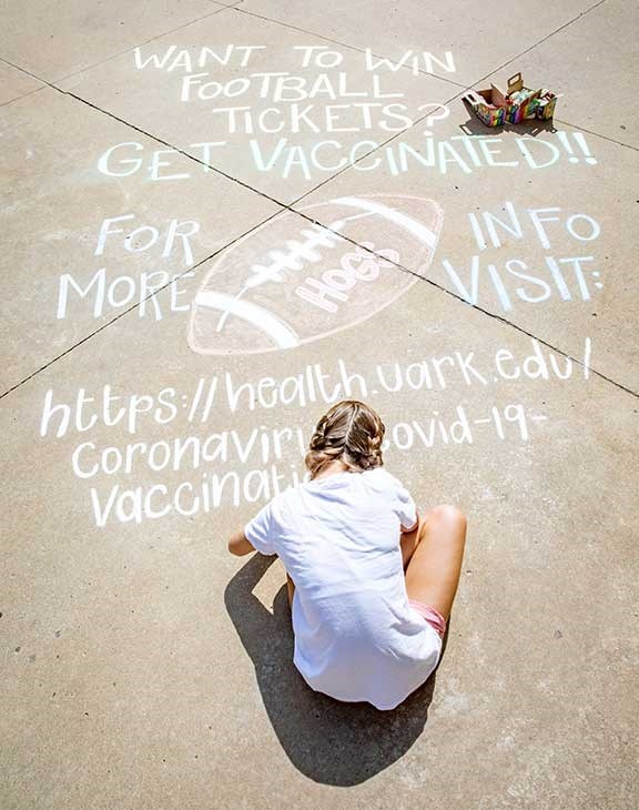 Studio art student Meredith Tinkle, sitting, helps create awareness of the university's vaccination incentive effort near the Arkansas Union.