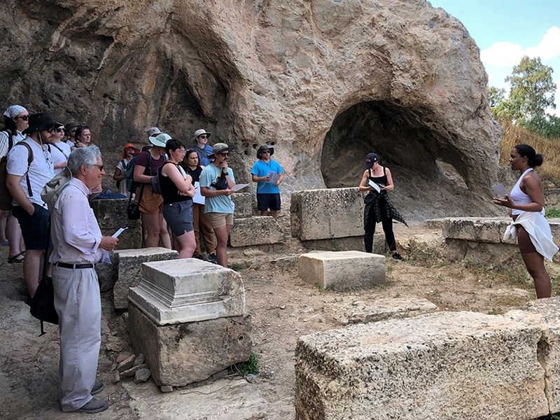 Daniel Levine (left) and participants of the 2022 Thanatopsis Summer Seminar listen to a site report by seminar member Trinity Rosa (University of Illinois at Urbana Champaign) at the Ploutonion at Eleusis.